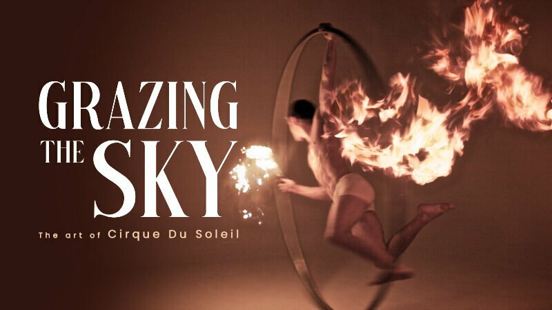 A cirque du soleil performer hangs from a ring that has been set on fire as cover art for Grazing The Sky