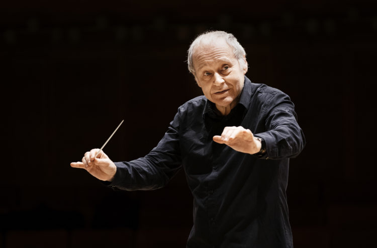 Conductor Adam Fischer stands holding a baton in his right hand wearing all black
