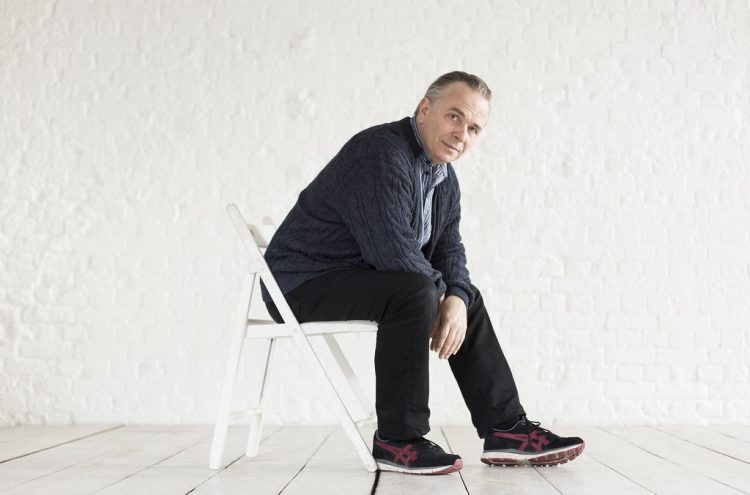 Mark Elder, Principal Artist at the OAE poses sitting in a chair leaning over his right knee and looking at the camera dressed in a casual cardigan trousers and trainers