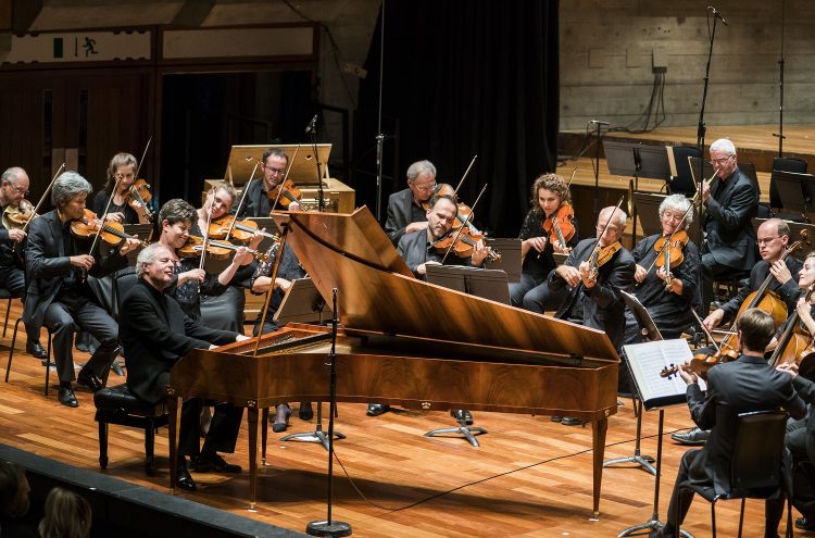 Sir Andras Schiff plays a piano forte on stage in front of an orchestra