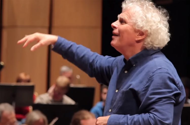 Conductor Sir Simon Rattle stands in rehearsal leading an orchestra dressed in a blue button up shirt