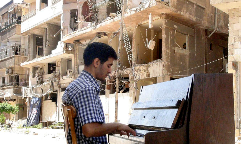 Pianist Aeham Ahmad sits playing an upright piano in front of destroyed buildings