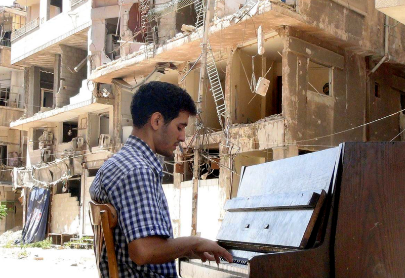 Pianist Aeham Ahmad sits playing an upright piano in front of destroyed buildings