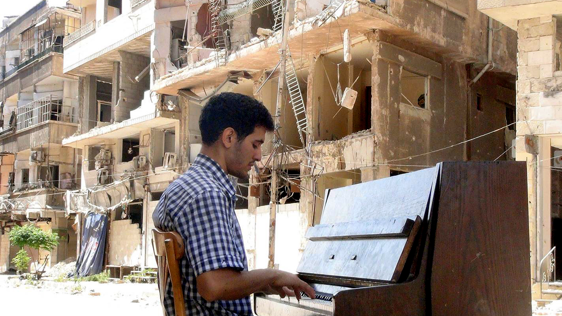 Pianist Aeham Ahmad sits playing an upright piano in front of destroyed buildings
