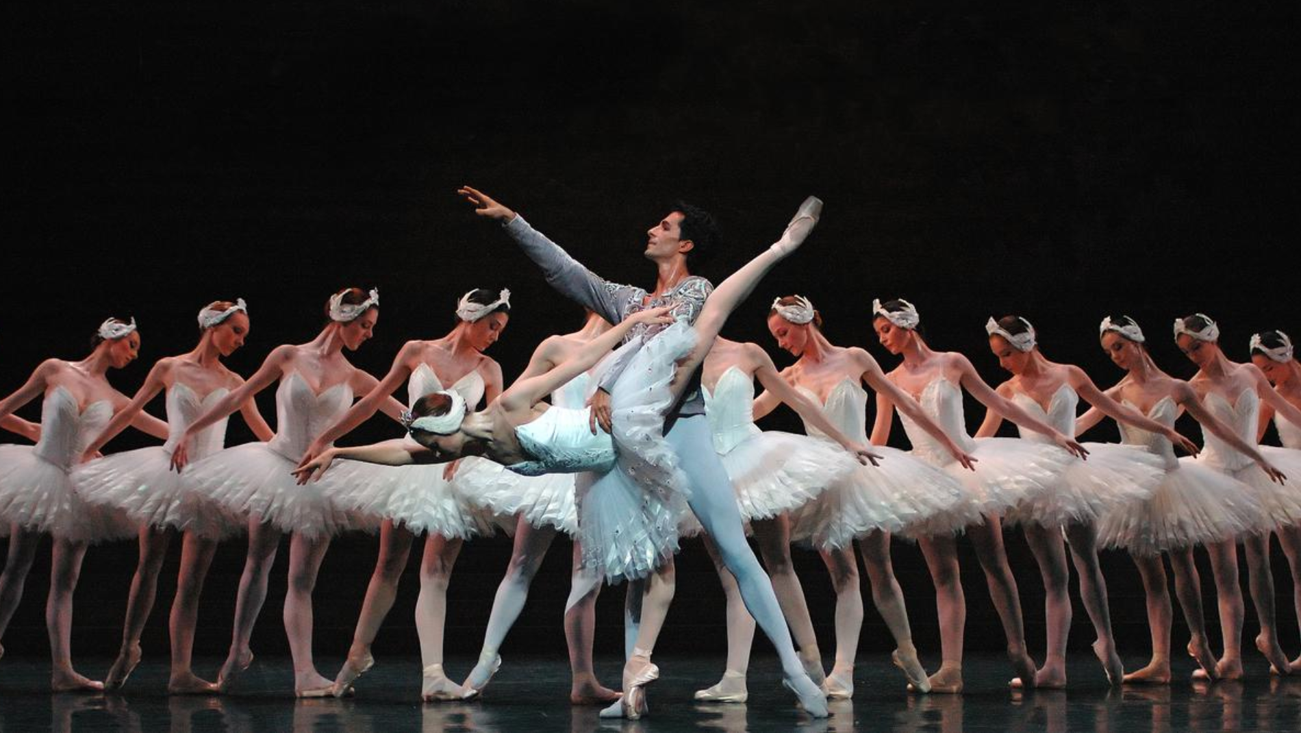 A female ballet dancer dressed in a traditional white Swan Lake costume poses on one leg with her arm outstretched. She is help by a male dancer who is dressed as the prince, he points his right arm out into the air. They stand in front of a row of female dancers who are all dressed in matching white swan lake costumes.