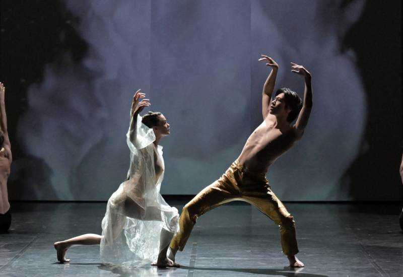 A male and female contemporary dancer stand at the center of the stage with their arms raised leaning towards eachother.