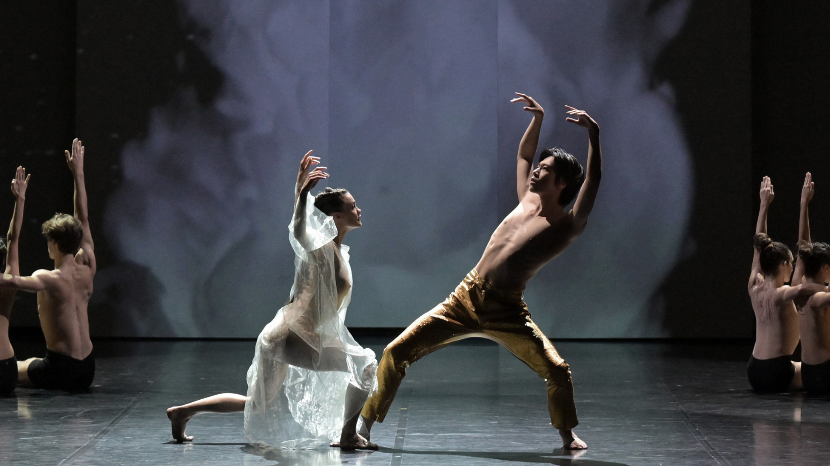 A male and female contemporary dancer stand at the center of the stage with their arms raised leaning towards eachother.