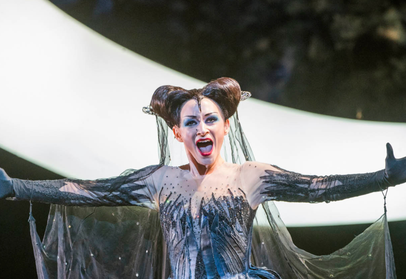 Sorpano Sabine Devielhe is dressed as The Queen of The Night costume featuring a medieval inspired black wig, and a see through and black dress which resemples black shards of the night. She stands singing with her mouth wide open