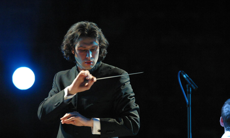 Conductor Vladimir Jurowski looks down at his music while conducting an orchestra, his hands are crossed in one hand he holds his conductor's batton. He is wearing a traditional suit and the background is black except for a single spotlight