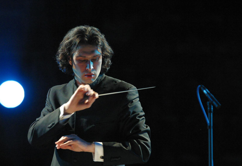 Conductor Vladimir Jurowski looks down at his music while conducting an orchestra, his hands are crossed in one hand he holds his conductor's batton. He is wearing a traditional suit and the background is black except for a single spotlight