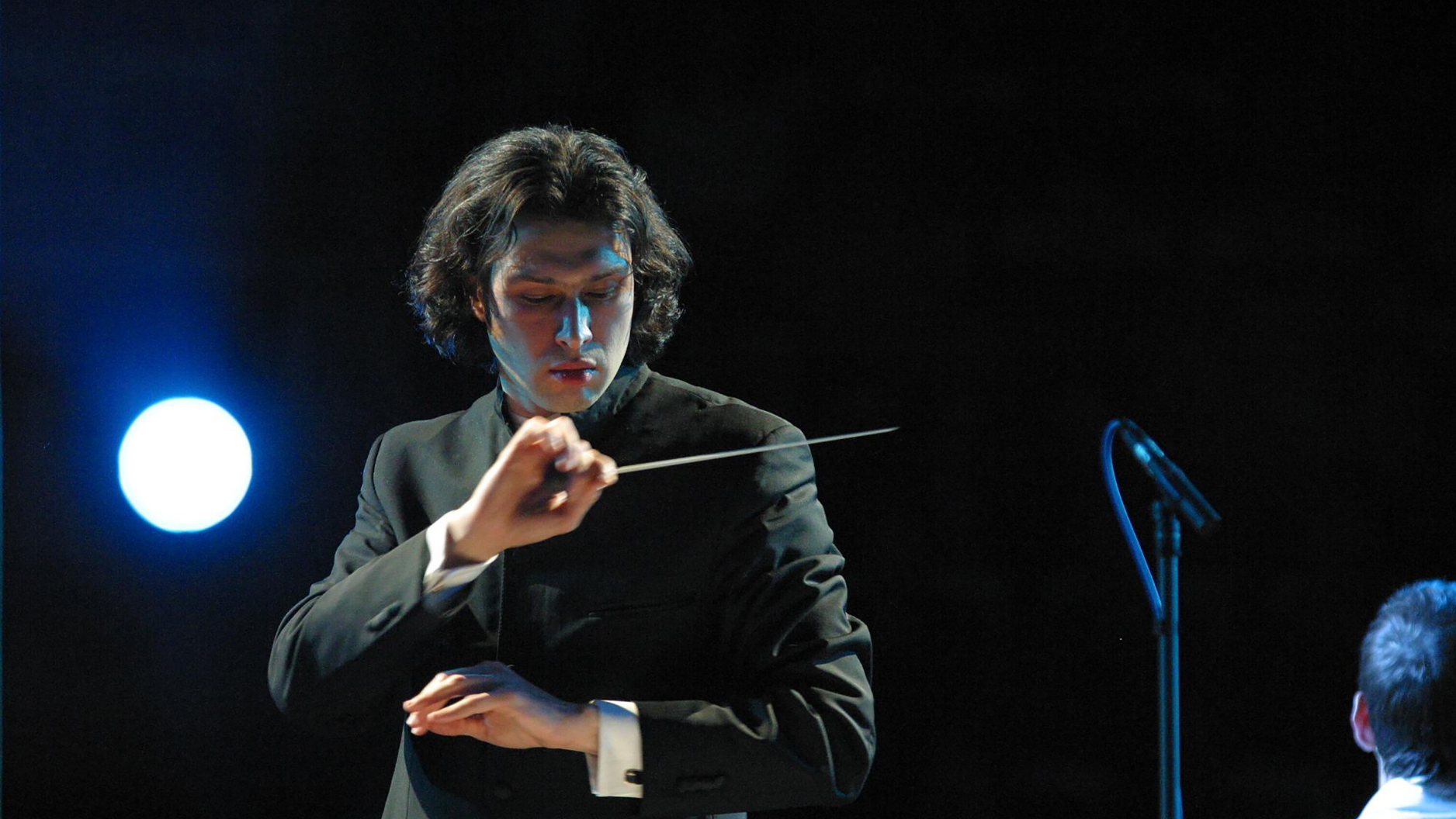 Conductor Vladimir Jurowski looks down at his music while conducting an orchestra, his hands are crossed in one hand he holds his conductor's batton. He is wearing a traditional suit and the background is black except for a single spotlight