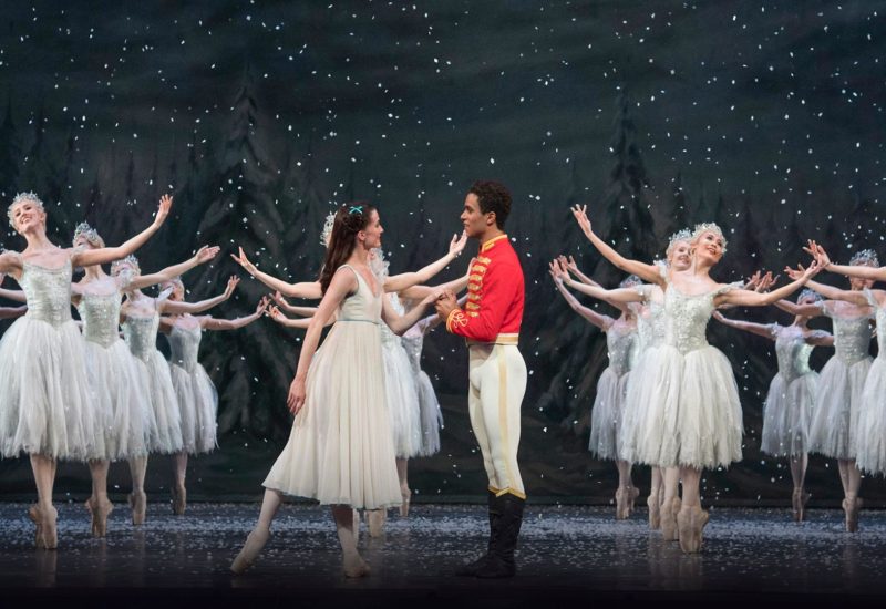 From the Royal Ballet's production of The Nutcracker. Clara dressed in a white flowing gown stands across from the Nutcracker Prince dressed in his red uniform. Behind them ballerinas dressed as snowflakes stand in formation.