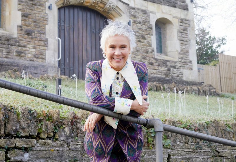 Director Emma Rice leans over a railing in front of a grey stone church. She is smiling and wearing a purple chevron suit with a long pointed white collar.