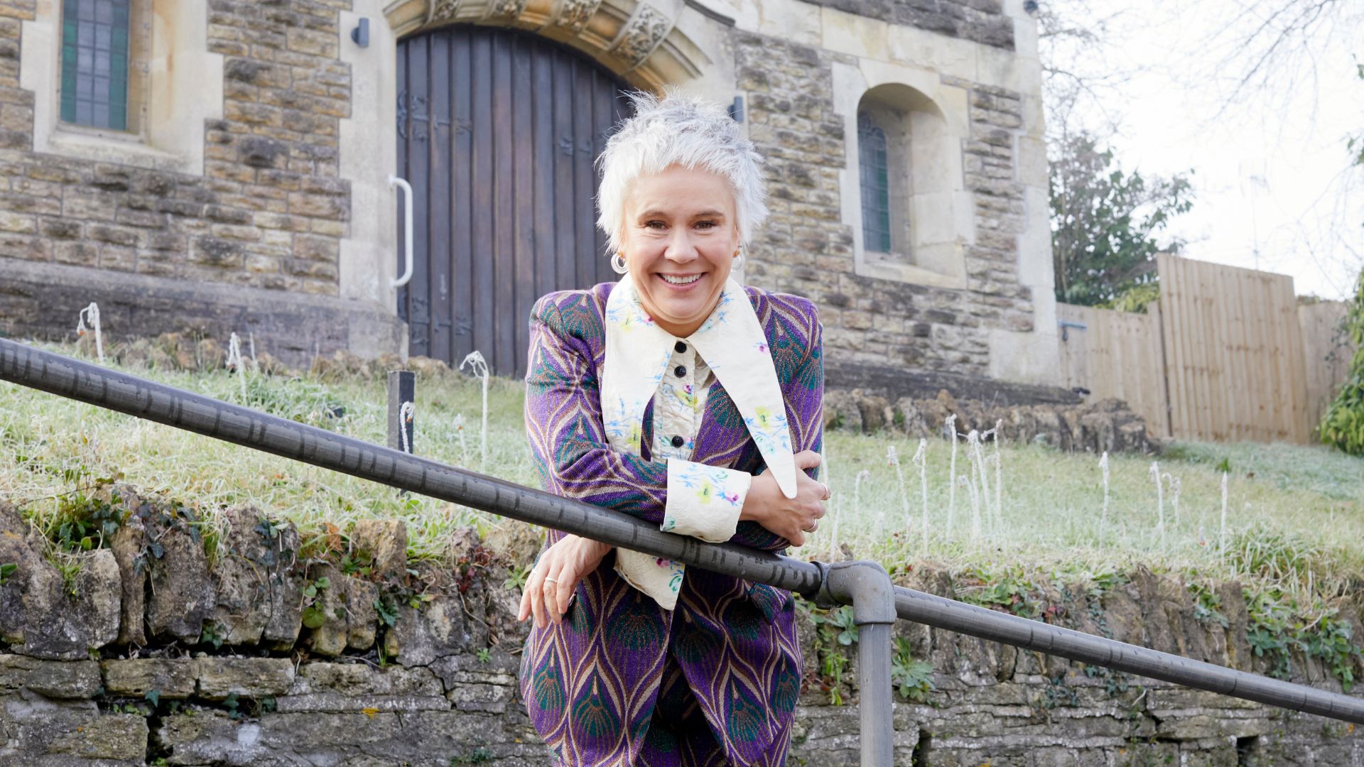 Director Emma Rice leans over a railing in front of a grey stone church. She is smiling and wearing a purple chevron suit with a long pointed white collar.