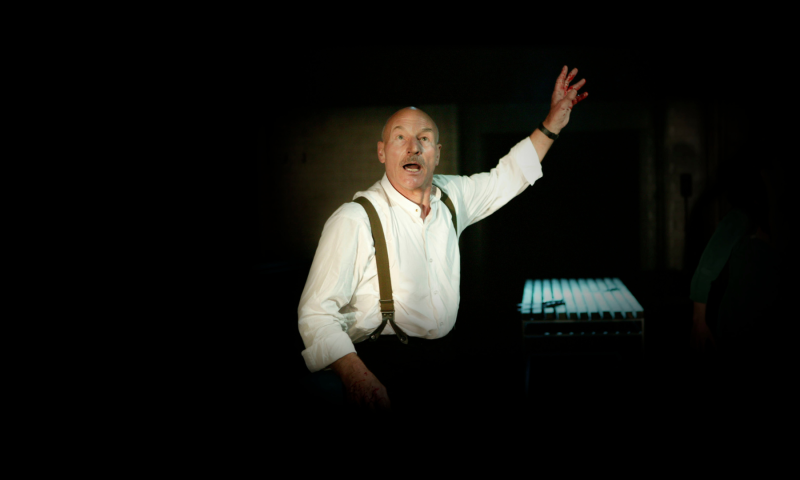 Sir Patrick Steward dressed in a white formal shirt with suspenders stands with his mouth open and one of his hands raised.