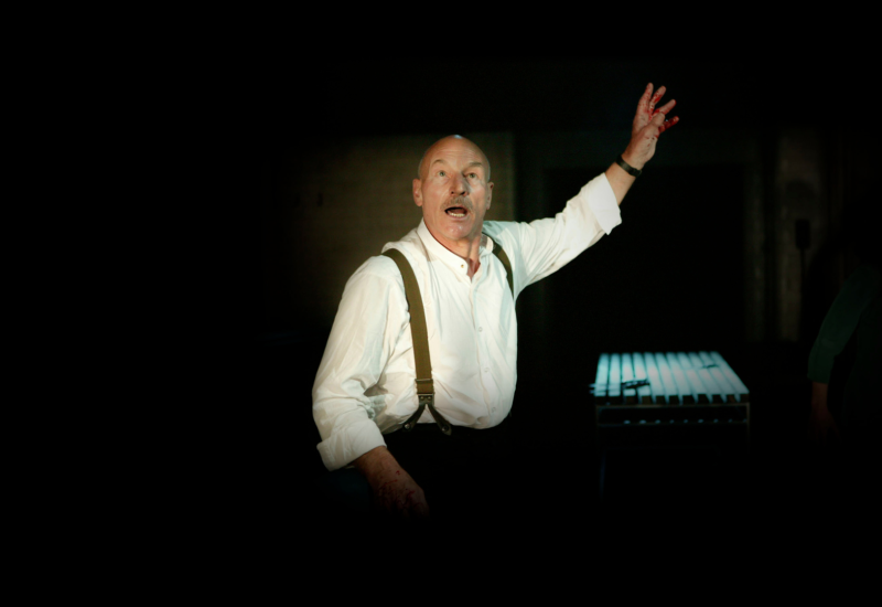 Sir Patrick Steward dressed in a white formal shirt with suspenders stands with his mouth open and one of his hands raised.