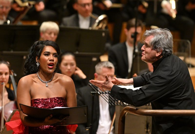 Sir Antonio Pappano leans over his conducting podium to the left to give a cue to the singer standing at his side. She is dressed in a red sequin dress and holding her music folder while singing.