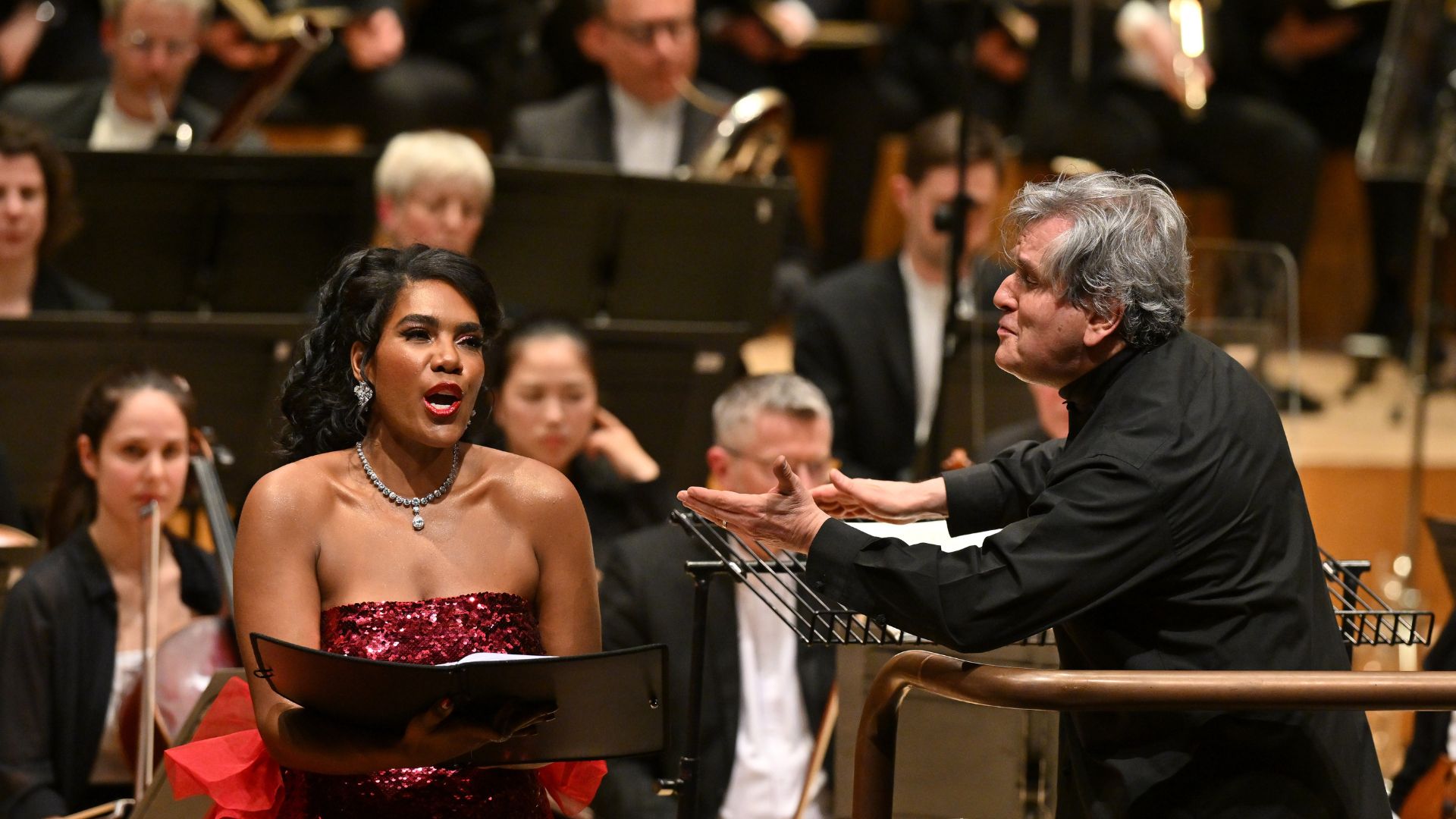 Sir Antonio Pappano leans over his conducting podium to the left to give a cue to the singer standing at his side. She is dressed in a red sequin dress and holding her music folder while singing.