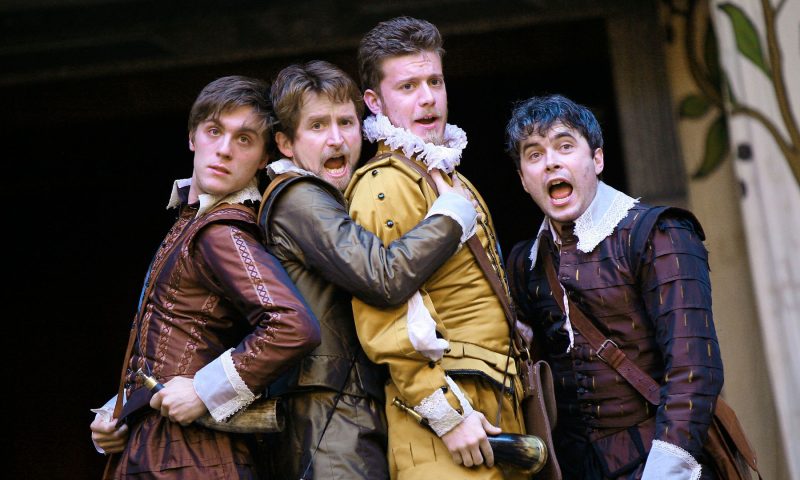 Four men dressed in Elizabethan clothing, with ruffed collars, pose together at the center of the stage looking shocked. From the Globe's production of Love's Labour's Lost.