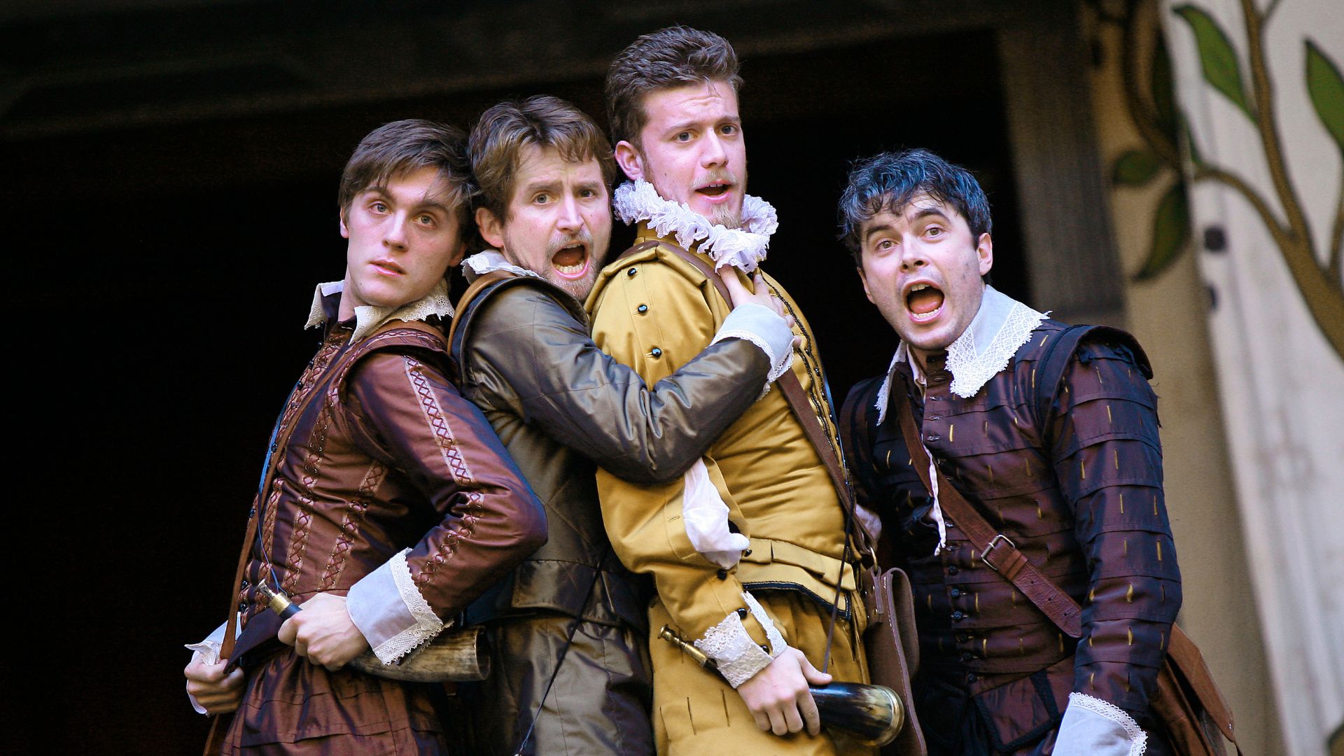 Four men dressed in Elizabethan clothing, with ruffed collars, pose together at the center of the stage looking shocked. From the Globe's production of Love's Labour's Lost.