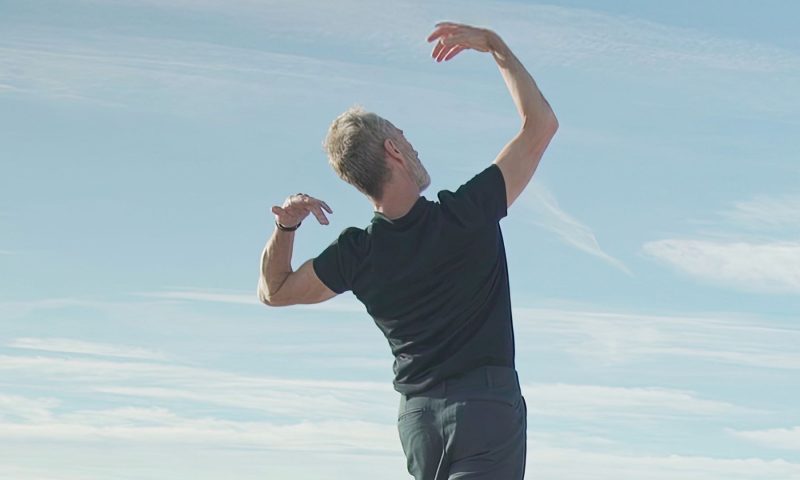 Choreographer Angelin Preljocaj stands on a rooftop with his back turned to the camera. His arms are above his head mid dance movement.