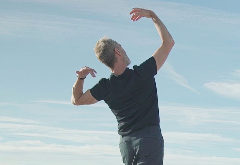 Choreographer Angelin Preljocaj stands on a rooftop with his back turned to the camera. His arms are above his head mid dance movement.