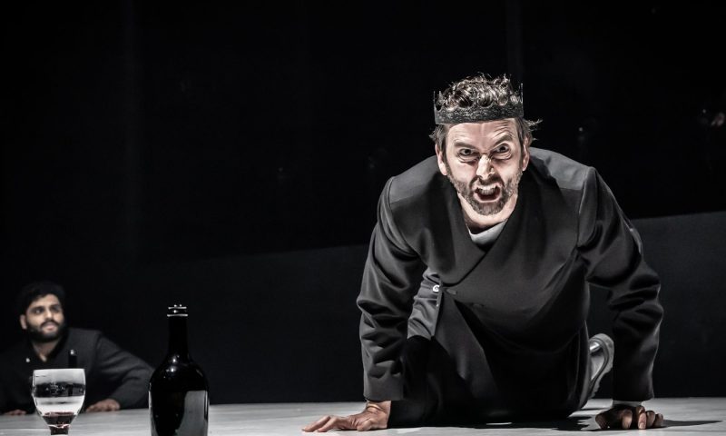 David Tennant dressed as Macbeth leans over a table facing the camera. His face is twisted in anger.