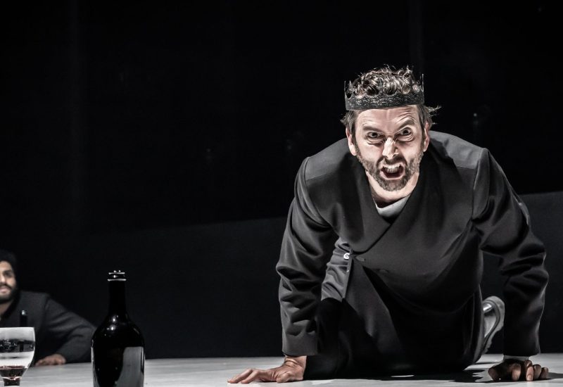 David Tennant dressed as Macbeth leans over a table facing the camera. His face is twisted in anger.