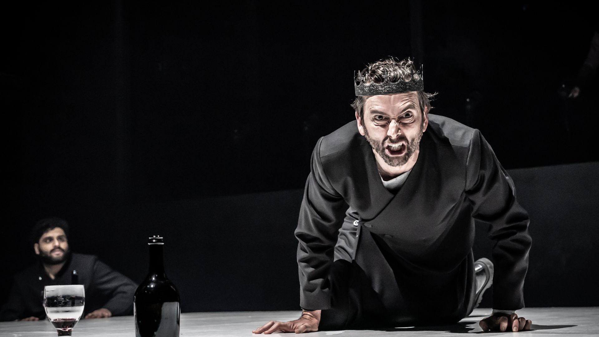 David Tennant dressed as Macbeth leans over a table facing the camera. His face is twisted in anger.