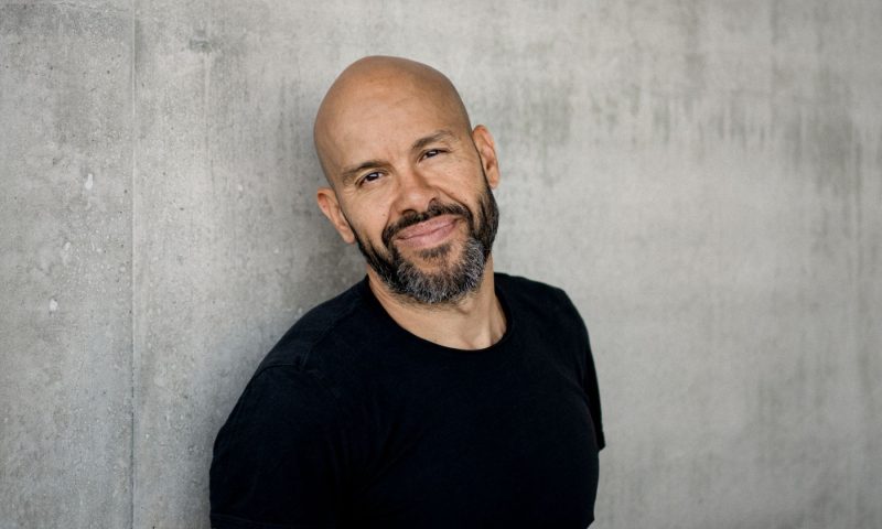 Choreographer Mourad Merzouki stands against a concrete wall in a black T shirt looking directly at the camera. He is only shown from the waist up