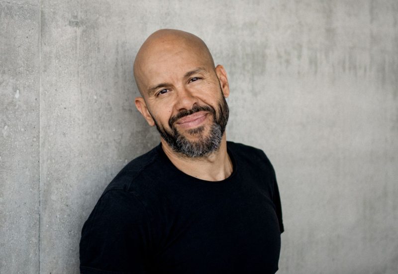 Choreographer Mourad Merzouki stands against a concrete wall in a black T shirt looking directly at the camera. He is only shown from the waist up
