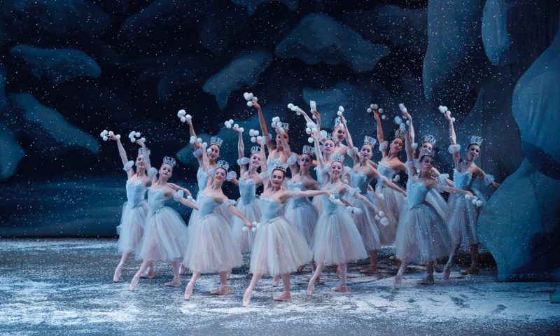 a group of ballet dancers dressed as Snow Flakes stand in a cluster at the right side of the stage. Their hands are raised and they hold clusters of snow balls in their hands. The stage is covered in snow.