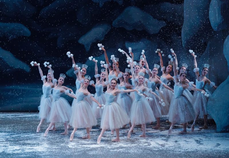 a group of ballet dancers dressed as Snow Flakes stand in a cluster at the right side of the stage. Their hands are raised and they hold clusters of snow balls in their hands. The stage is covered in snow.