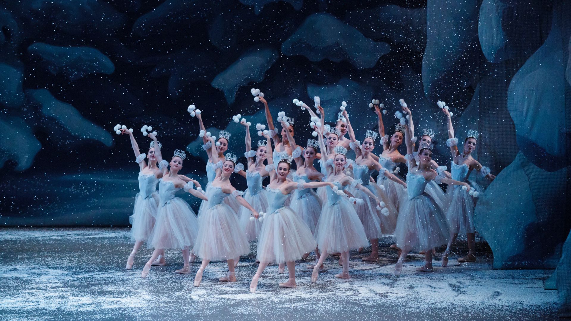 a group of ballet dancers dressed as Snow Flakes stand in a cluster at the right side of the stage. Their hands are raised and they hold clusters of snow balls in their hands. The stage is covered in snow.
